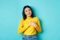 Portrait of beautiful heartfelt woman holding hands on heart, smiling and listening compassionate, standing over blue