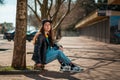 Portrait of a beautiful hipster teen girl in a leather jacket sitting on a skateboard. Side view. There`s an alley with trees in Royalty Free Stock Photo