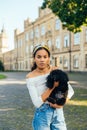 Portrait of a beautiful hispanic woman with a cute black dog in her arms, standing on the street and looking at the camera Royalty Free Stock Photo