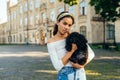 Portrait of a beautiful hispanic woman with a cute black dog in her arms, standing on the street and looking at the camera Royalty Free Stock Photo