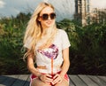 Portrait beautiful long-haired blond girl in vintage sunglasses, T-shirt and shorts sitting on a bench with a cool drink on a hot Royalty Free Stock Photo