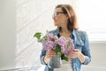 Portrait of beautiful mature woman at home with bouquet of lilac flowers. Background of home interior dining near the window