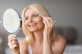 Portrait of beautiful middle aged blonde woman applying eye-shadows and looking in mirror, sitting in bedroom Royalty Free Stock Photo