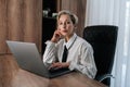 Portrait of beautiful middle-aged businesswoman sitting on desk at office working with laptop, looking confident at the Royalty Free Stock Photo