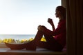 Portrait of a beautiful modern young woman sitting on a window sill with a mug, and enjoying a sea view. Royalty Free Stock Photo