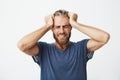 Portrait of beautiful nordic guy with trendy haircut and beard holding head with hands suffering from headache after a