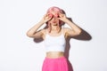 Portrait of beautiful party girl in pink wig, holding macaroons over eyes and pouting silly, standing over white Royalty Free Stock Photo