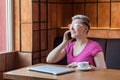 Portrait of beautiful satisfied happy young bussineswoman with blonde short hair in pink t-shirt is sitting in cafe, talking on