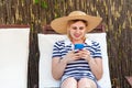 Portrait of beautiful satisfied young adult blogger woman in hat and dress is sitting on sunbed and holding phone, looking at Royalty Free Stock Photo