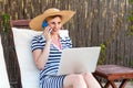 Portrait of beautiful satisfied young adult businesswoman in hat and dress sitting on sunbed with laptop and working online, Royalty Free Stock Photo