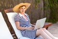 Portrait of beautiful satisfied young adult freelancer woman in hat and dress is sitting on cozy sunbed with laptop and working Royalty Free Stock Photo