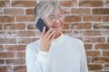 Portrait of beautiful senior woman white haired using phone standing against a brick wall smiling Royalty Free Stock Photo