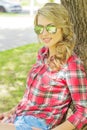 Portrait of a beautiful girl with large plump lips curls in denim shorts and a shirt in sunglasses sitting near a tree Royalty Free Stock Photo