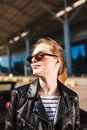 Portrait of beautiful smiling girl in black leather jacket and sunglesses dreamily looking aside with airport on