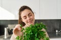 Portrait of beautiful smiling girl with bouquet of parsley, standing in the kitchen and cooking, adding herbs to healthy