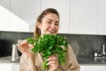 Portrait of beautiful smiling girl with bouquet of parsley, standing in the kitchen and cooking, adding herbs to healthy