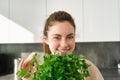 Portrait of beautiful smiling girl with bouquet of parsley, standing in the kitchen and cooking, adding herbs to healthy