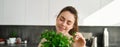 Portrait of beautiful smiling girl with bouquet of parsley, standing in the kitchen and cooking, adding herbs to healthy