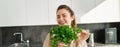 Portrait of beautiful smiling girl with bouquet of parsley, standing in the kitchen and cooking, adding herbs to healthy