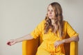 Portrait of beautiful smiling young blonde woman with long curly hair in retro yellow dress sitting in armchair Royalty Free Stock Photo