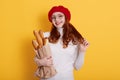 Portrait of beautiful smiling young woman with red hair, holding paper bag with bread baguette, stands over yellow background, Royalty Free Stock Photo