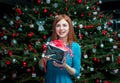 Portrait of beautiful smiling young woman, standing near Christmas tree, holding gift Royalty Free Stock Photo