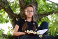 Portrait of beautiful teenage girl with glasses, sitting on branch of large tree, an oak in summer park with book in her hands. Royalty Free Stock Photo