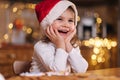 Portrait of beautiful tree year girl in santa hat sitting by the table in front of kitchen decorated fairy lights