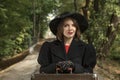 Portrait of beautiful woman in black hat with brims, classic jaket and gloves, hands on old suitcase. Forest background