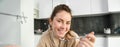 Portrait of beautiful woman at home, eating her breakfast, holding spoon, leaning on worktop and having cereals with Royalty Free Stock Photo