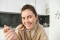 Portrait of beautiful woman at home, eating her breakfast, holding spoon, leaning on worktop and having cereals with Royalty Free Stock Photo