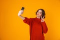 Portrait of beautiful woman holding and taking selfie photo on cell phone while standing isolated over yellow background Royalty Free Stock Photo
