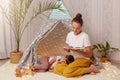 Portrait of beautiful young adult mother holding book and reading fairy tale for her little daughter, kid laying on floor and Royalty Free Stock Photo