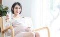 Portrait beautiful young asian pregnant woman hand holding drink milk sit on dofa with big window in livingroom. health care and Royalty Free Stock Photo