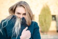 Portrait of beautiful young blonde girl in blue coat, who froze and hides in her collar, at sunset, against brick wall