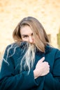 Portrait of beautiful young blonde girl in blue coat, who froze and hides in her collar, at sunset, against brick wall