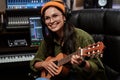 Portrait of beautiful young brunette, female artist smiling aside, playing ukulele while sitting in recording studio Royalty Free Stock Photo