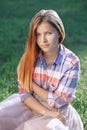 Portrait of beautiful young Caucasian woman with long red hair in plaid shirt and pink tutu tulle skirt, sitting on grass in park Royalty Free Stock Photo