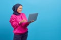 Portrait of beautiful young female college student with laptop smiling wearing pink jacket isolated on blue background