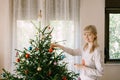 Portrait of a beautiful young girl decorating a lush Christmas tree with toys and balls in her apartment.