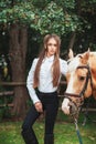 Portrait beautiful young girl in white shirt and black pants with beauty long hair next horse in forest. Fashionable elegance woma Royalty Free Stock Photo