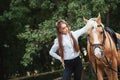 Portrait beautiful young girl in white shirt and black pants with beauty long hair next horse in forest. Fashionable elegance woma Royalty Free Stock Photo