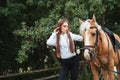 Portrait beautiful young girl in white shirt and black pants with beauty long hair next horse in forest. Fashionable elegance woma Royalty Free Stock Photo