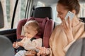 Portrait of beautiful young mother wearing beige sweater talking on the mobile phone in the car while the child is sitting next to Royalty Free Stock Photo