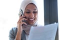 Beautiful young muslim business woman wearing hijab talking with her smart phone while holding papers looking through the window Royalty Free Stock Photo