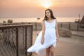 Portrait of beautiful young pregnant woman in white dress walking on wooden walkway near seacoast at sunset. Royalty Free Stock Photo