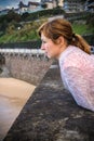 Portrait of beautiful young red haired woman leaning on the wall, observing waves and surfers on the shore of atlantic coast Royalty Free Stock Photo