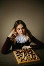 Portrait of a beautiful young woman in an antique dress with a white frill, who is sitting in front of a chess board and thinking Royalty Free Stock Photo