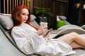 Portrait of beautiful young woman in bathrobe relaxing with cellphone lying on deckchair by poolside and drinking Royalty Free Stock Photo