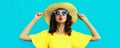 Portrait of beautiful young woman blowing her lips sending sweet air kiss posing wearing yellow summer straw hat, dress on blue Royalty Free Stock Photo